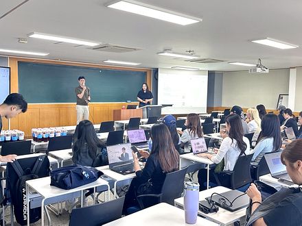 강의실에서 학생들이 노트북으로 디자인 툴을 실습하고 있고, 앞에서 강사가 설명하고 있습니다.